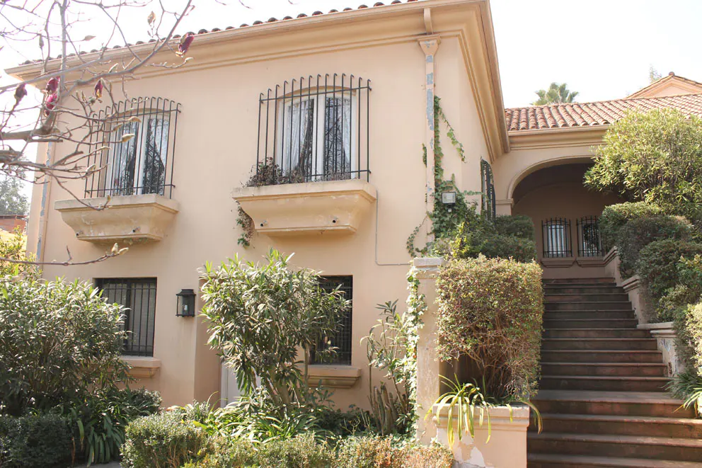Two-story beige house with black barred windows and a red tile roof. A stone staircase leads to an arched entryway. Green bushes surround the house.
