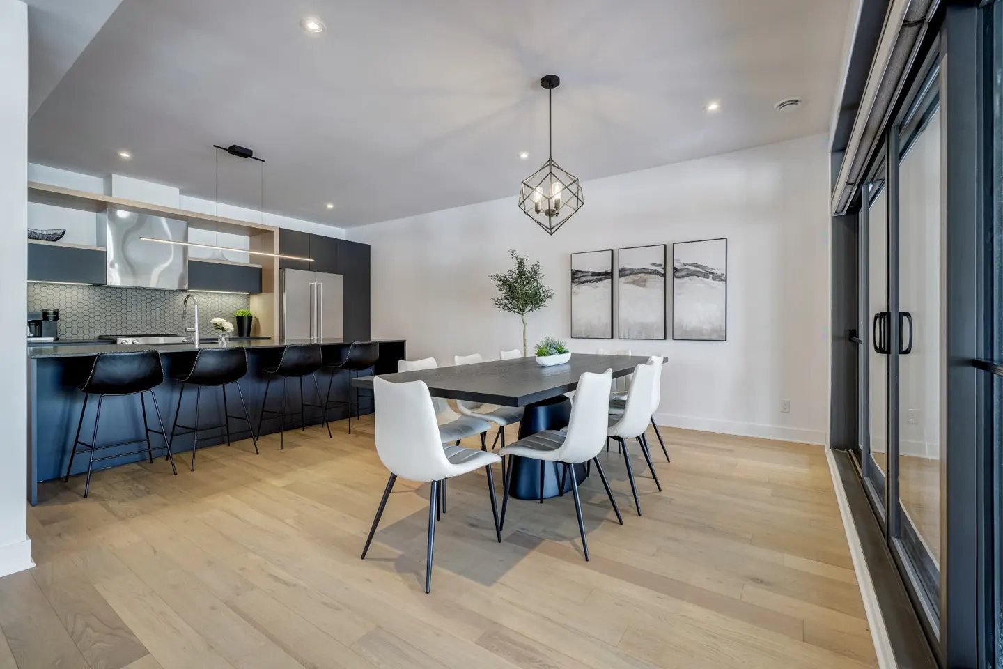 A modern kitchen and dining area with a black island, dining table, and white chairs on light wood floors. Artwork hangs on the white wall.