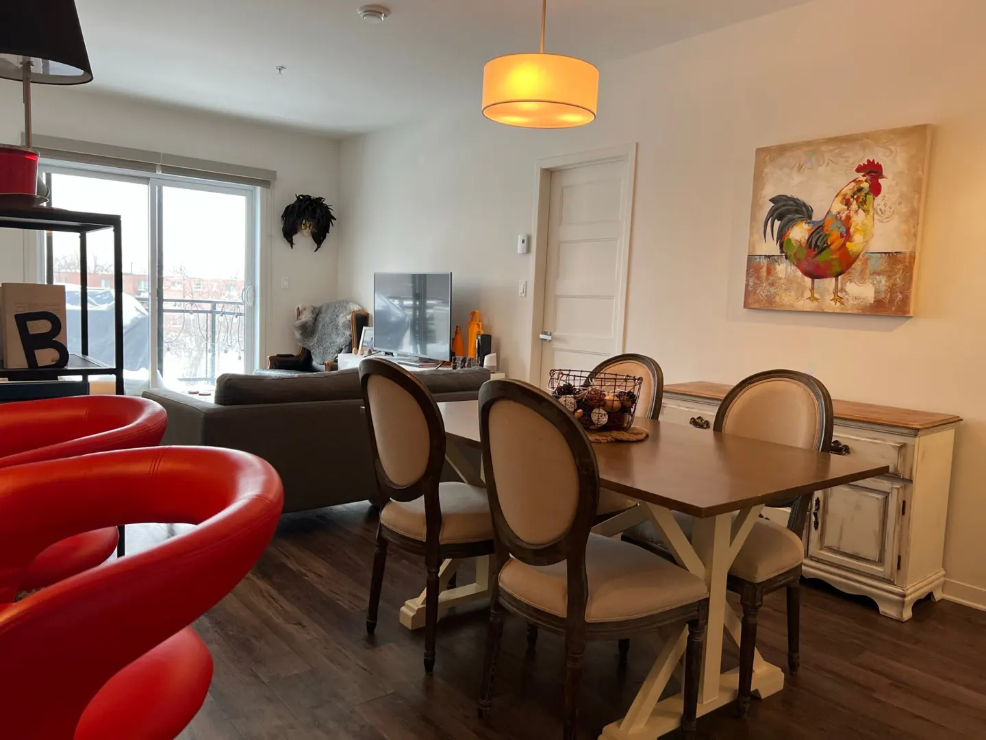 Open-concept living space with a dining table, red chairs, and a brown sofa. A rooster painting hangs above a white sideboard.