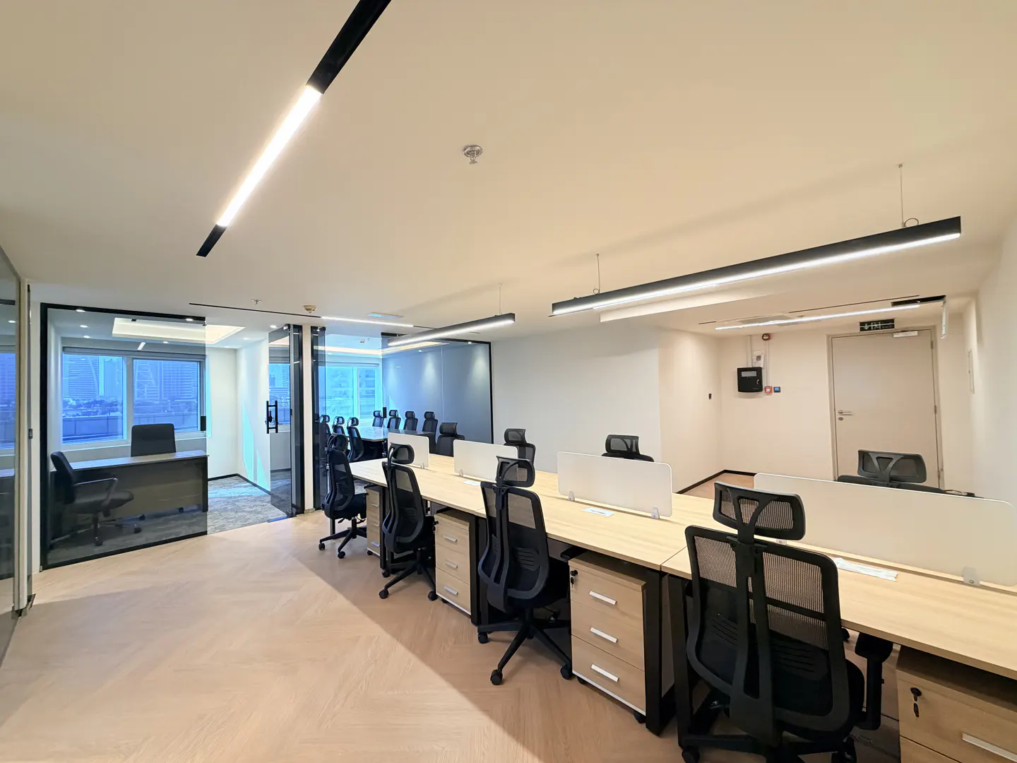 Bright, modern office space with rows of desks and black chairs. Glass-walled offices are visible in the background.