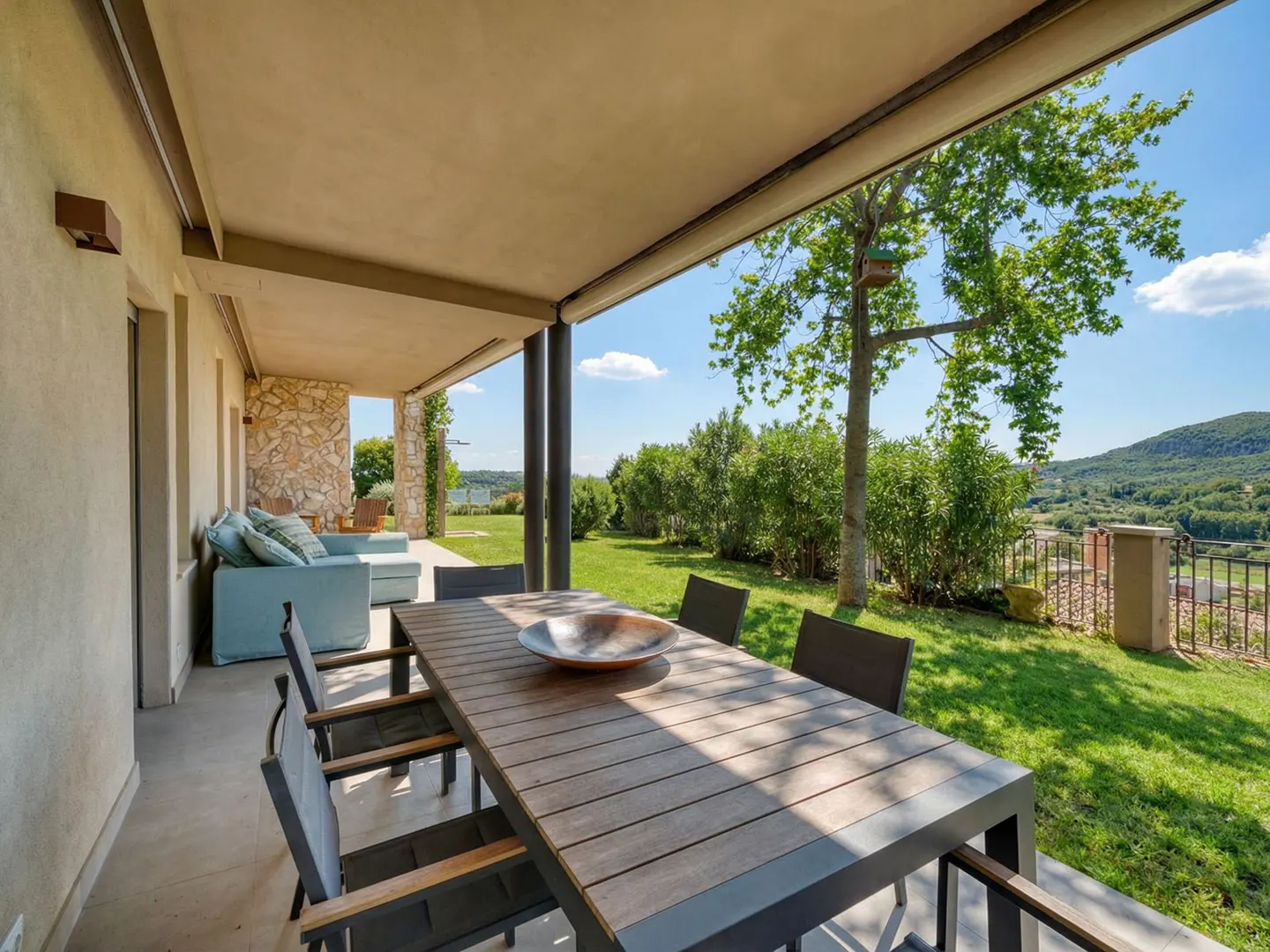 Covered patio with a long wooden table and chairs, a blue sofa, and a view of green grass, trees, and distant hills.