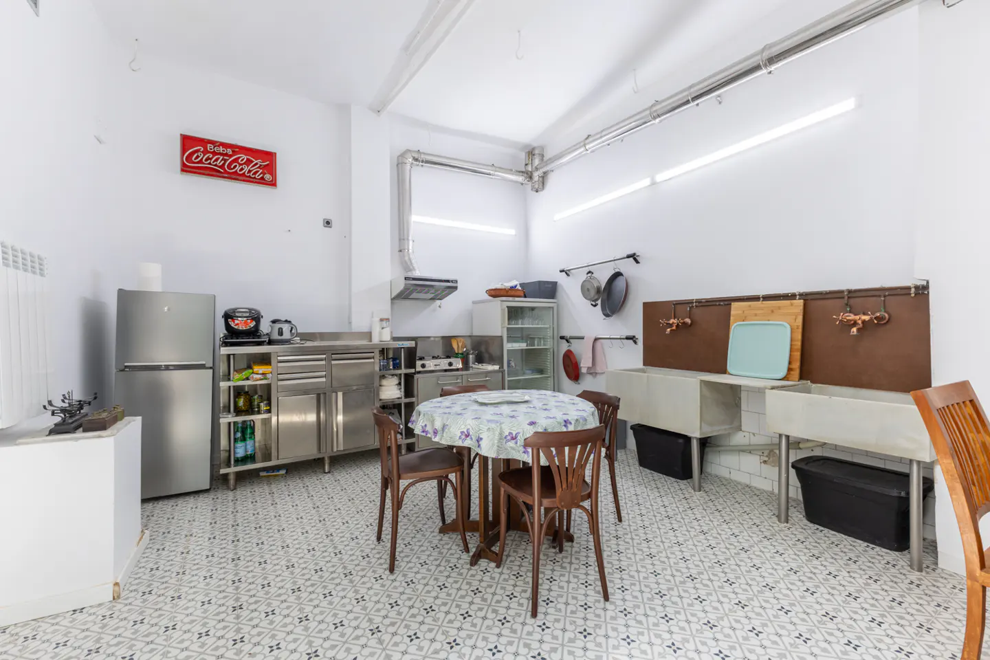 Bright kitchen with stainless steel appliances, white walls, and patterned tile floor. A round table with chairs sits in the center. A Coca-Cola sign hangs on the wall.