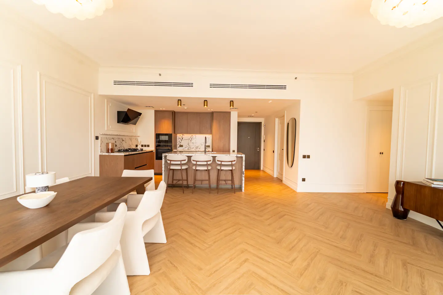 Bright, open-concept apartment with herringbone wood floors. Kitchen island with stools, dining table with white chairs, and modern decor.