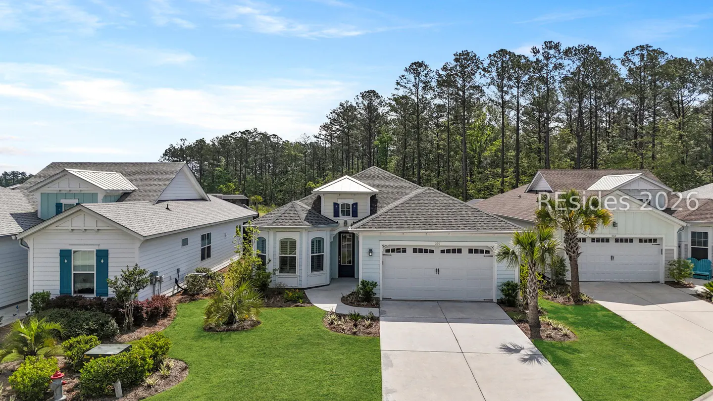 Aerial view of a light blue single-story house with a gray roof, white garage door, and green lawn. Trees in the background.