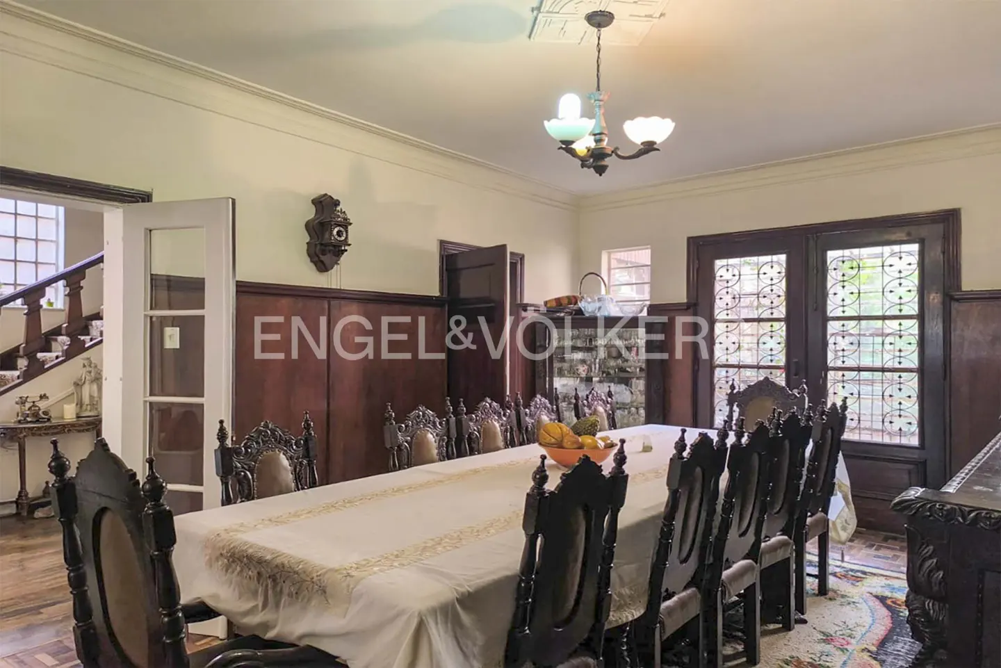 A formal dining room with a long table, ornate dark wood chairs, and a vintage chandelier. The Engel & Volkers logo is visible.