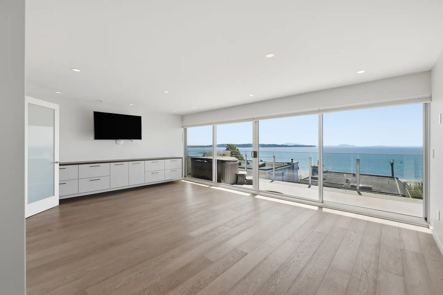 Modern living room with light wood floors, white cabinets, wall-mounted TV, and floor-to-ceiling windows overlooking the sea.