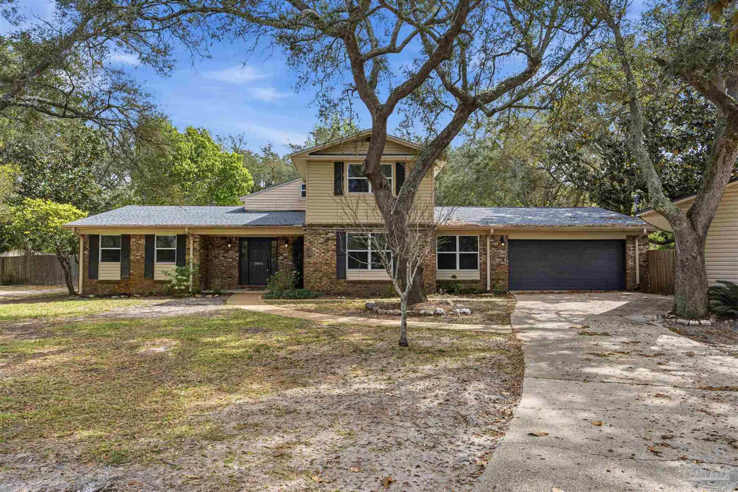 Two-story house with brick and beige siding, black shutters, and a dark gray garage door. Trees frame the house against a blue sky.
