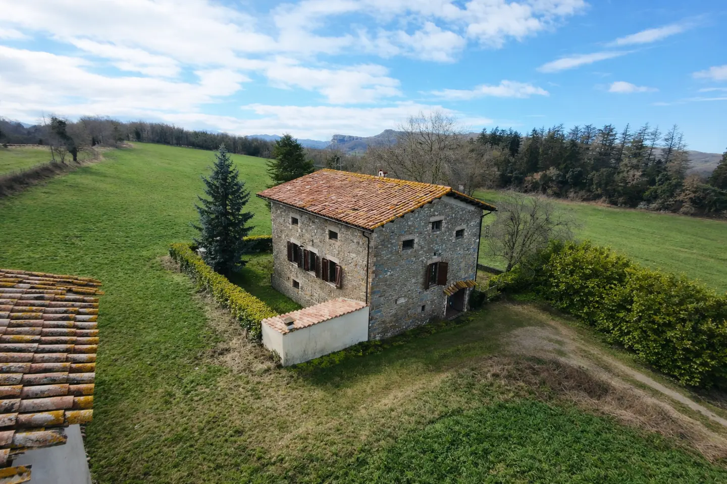 Stone house with a red tile roof, surrounded by green fields and trees under a blue sky with white clouds.