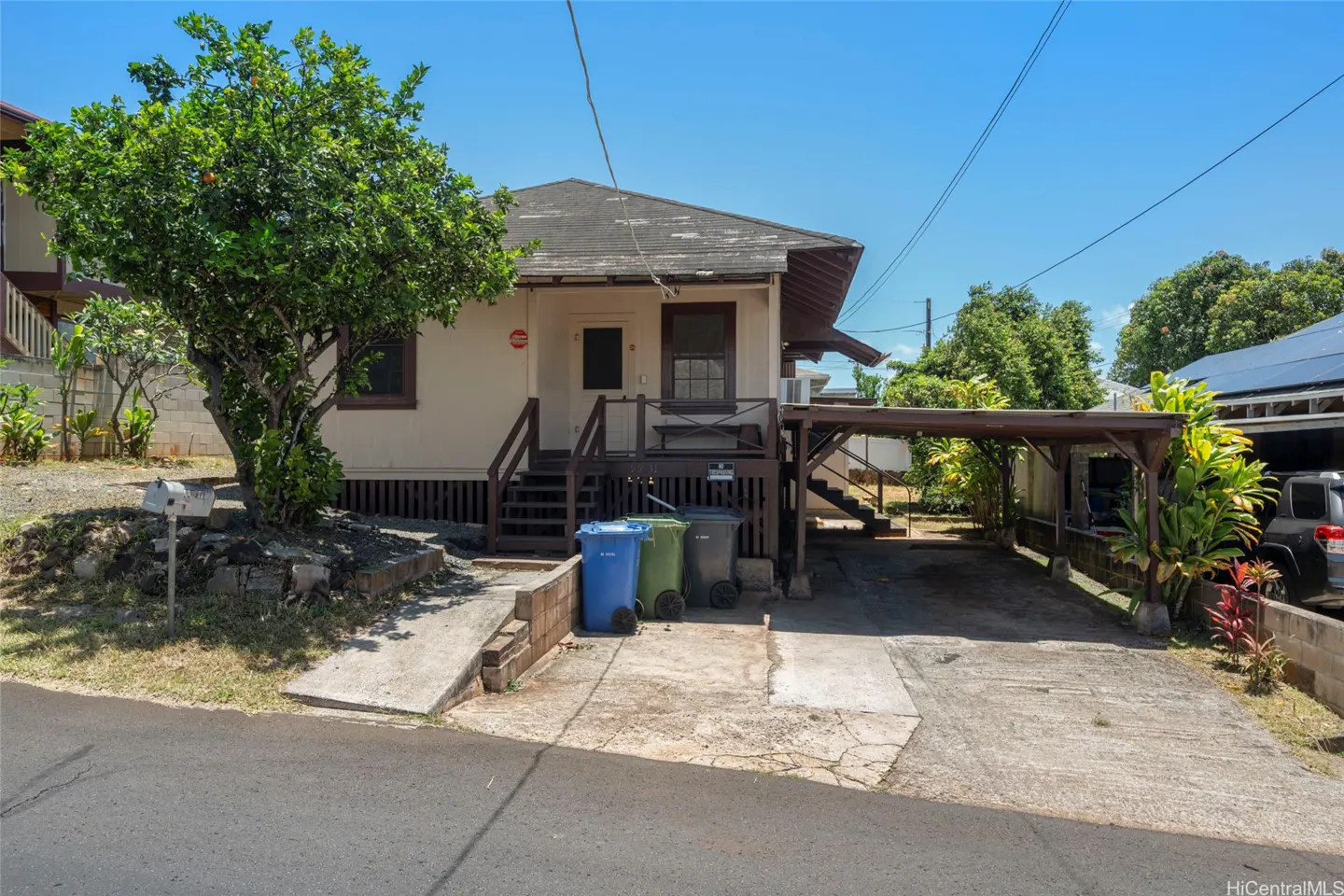 Exterior view of a single-story beige house with brown trim, a carport, and a green tree in the front yard. Three trash bins are near the stairs.