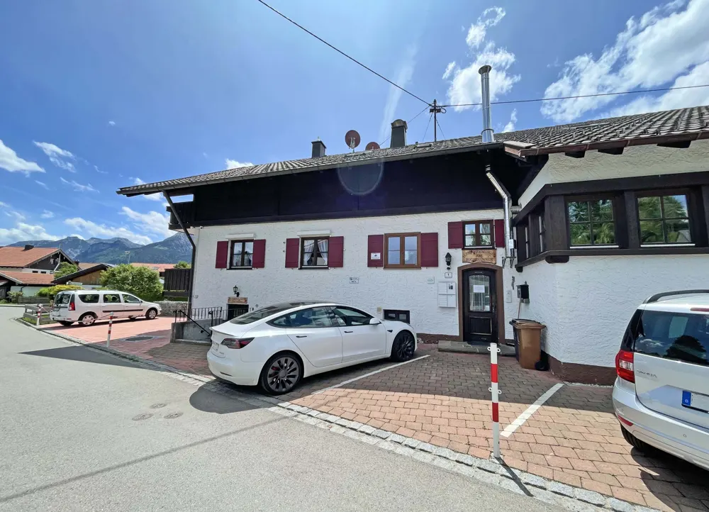 Exterior of a white building with red shutters, a white Tesla parked in front, and mountains in the background.