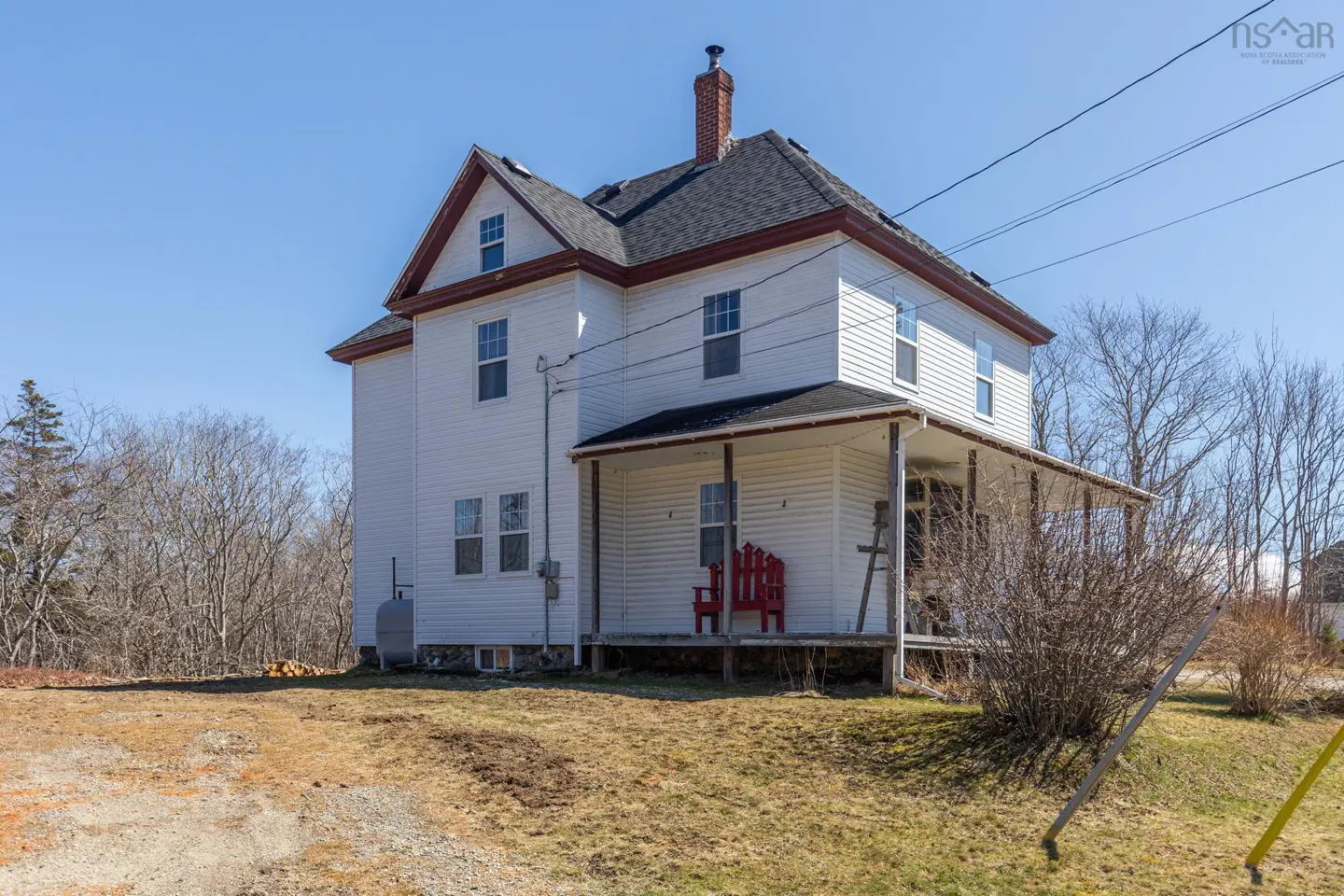 Two-story white house with a dark roof, red trim, and a covered porch with red chairs. Blue sky and trees in the background.
