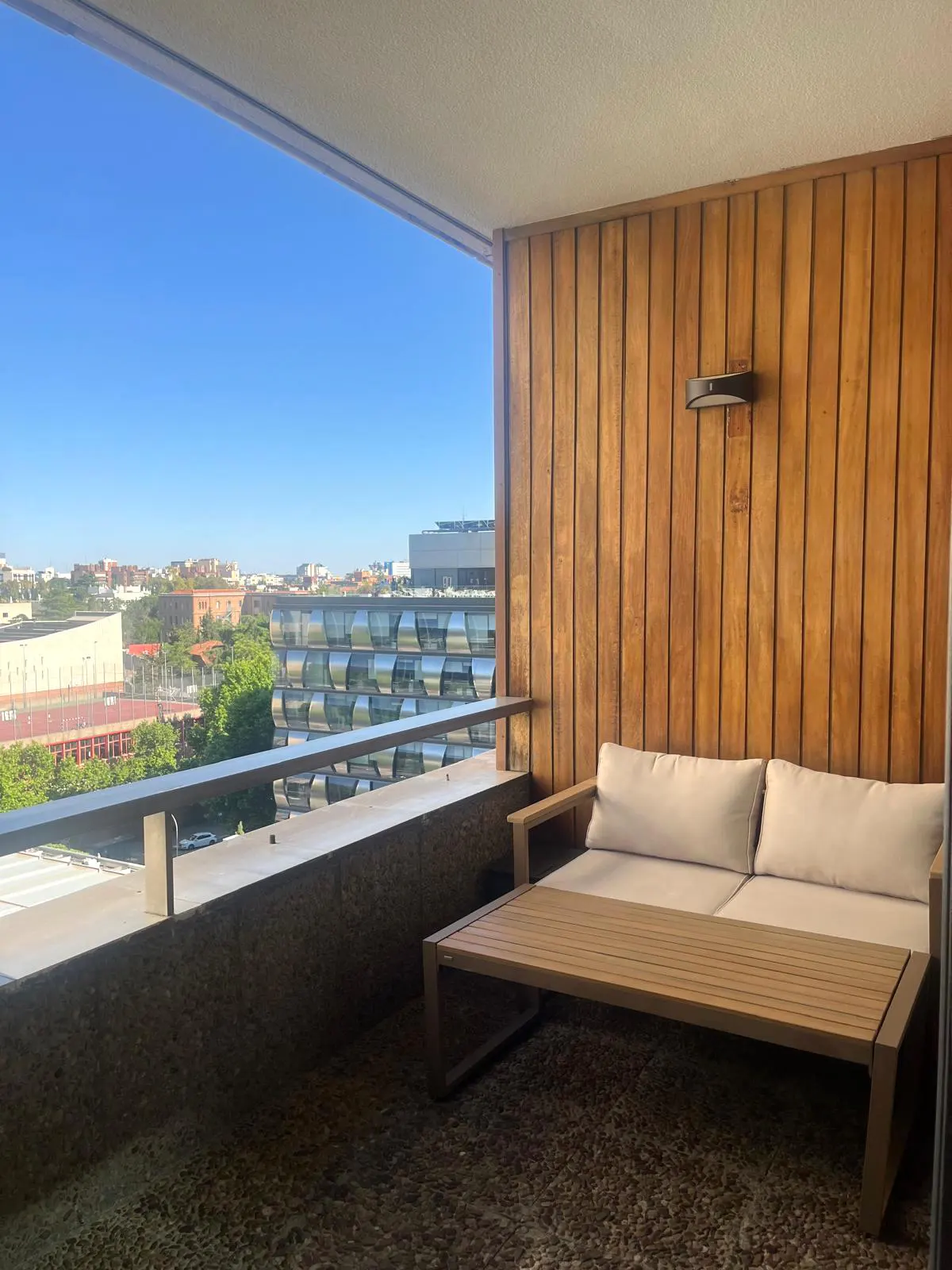 Balcony with a wooden wall, a light brown sofa, and a view of the city under a clear blue sky.