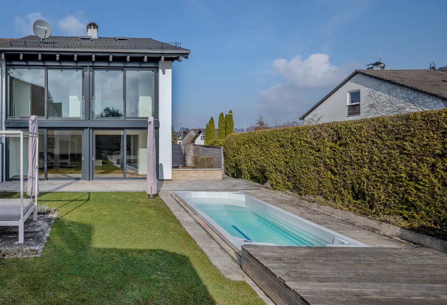Modern home exterior with a rectangular pool, green lawn, and large windows. A tall hedge provides privacy.