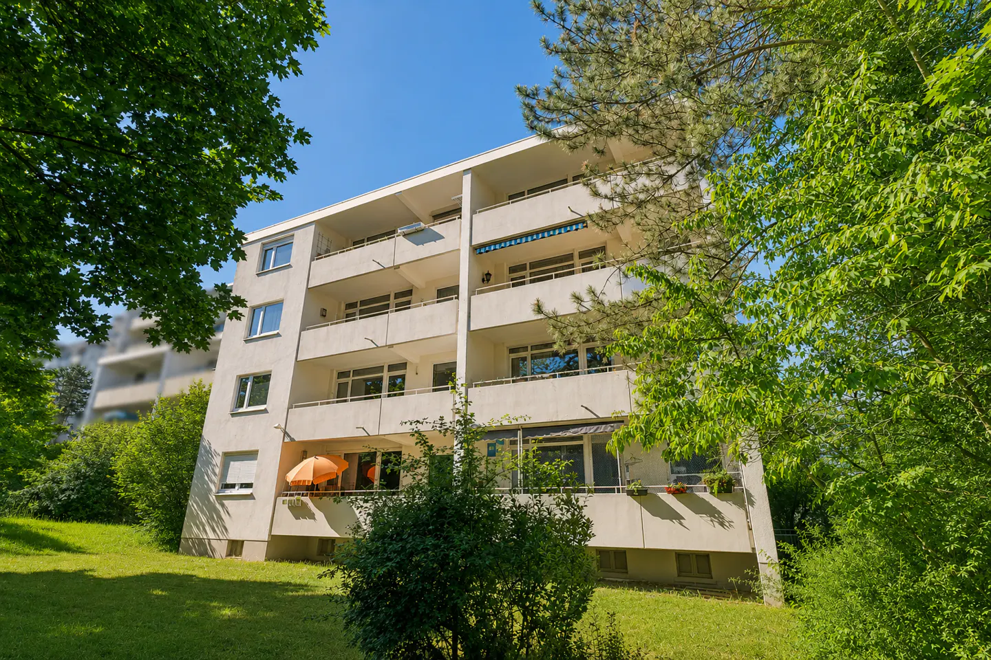 A four-story white apartment building with balconies, surrounded by lush green trees and a grassy lawn under a blue sky.