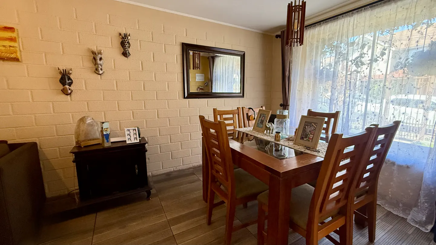 A dining room with a wooden table and chairs, a brick wall with animal masks, and a window with sheer curtains.