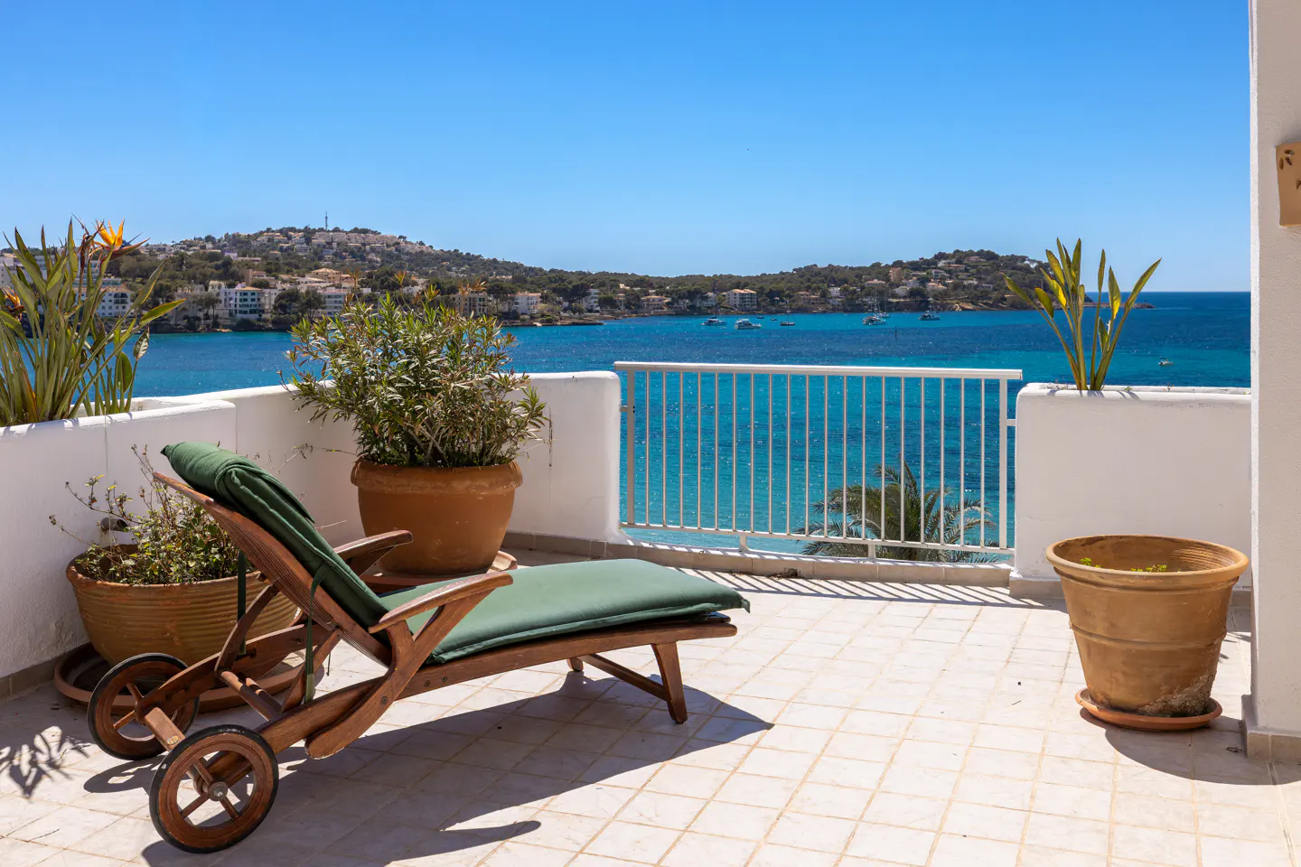 Balcony view with a wooden lounge chair, potted plants, and a turquoise ocean backdrop under a clear blue sky.