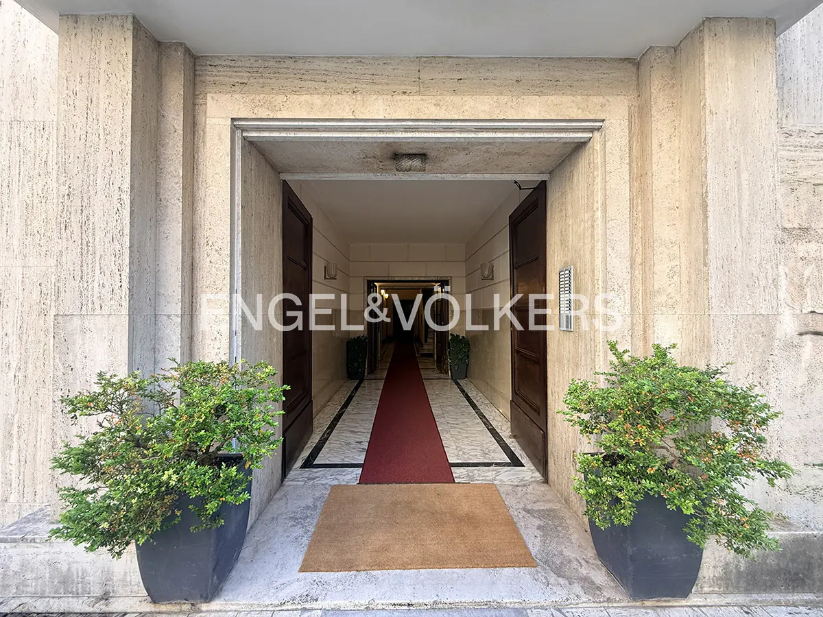 Entrance to a building with stone walls, dark wood doors, and potted plants. A red carpet runs down the hallway.