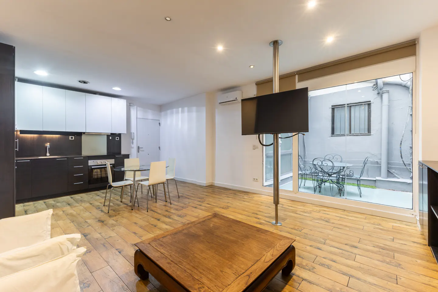 Bright apartment interior with wood floors, a kitchen, dining table, and a TV mounted on a pole. A patio is visible through a glass door.