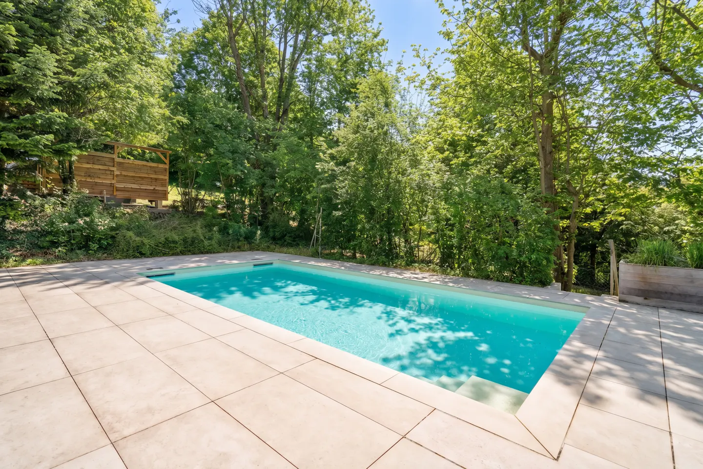 A rectangular turquoise pool is surrounded by light gray tiles and lush green trees.