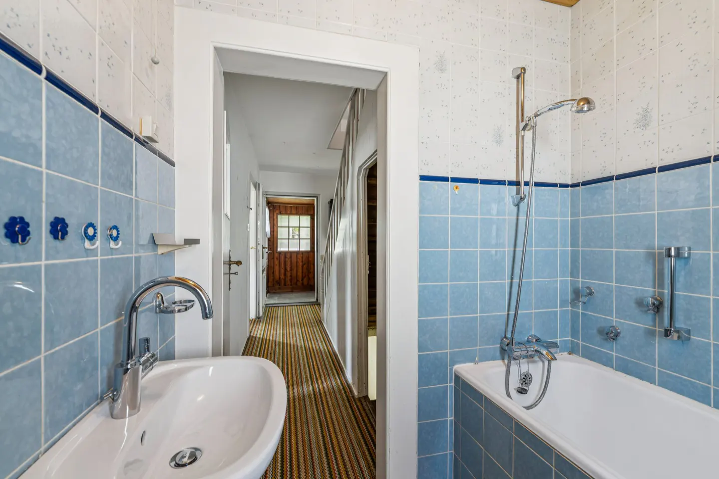 Bathroom with blue tile, white sink, and bathtub. A hallway with a wooden door is visible through the doorway.