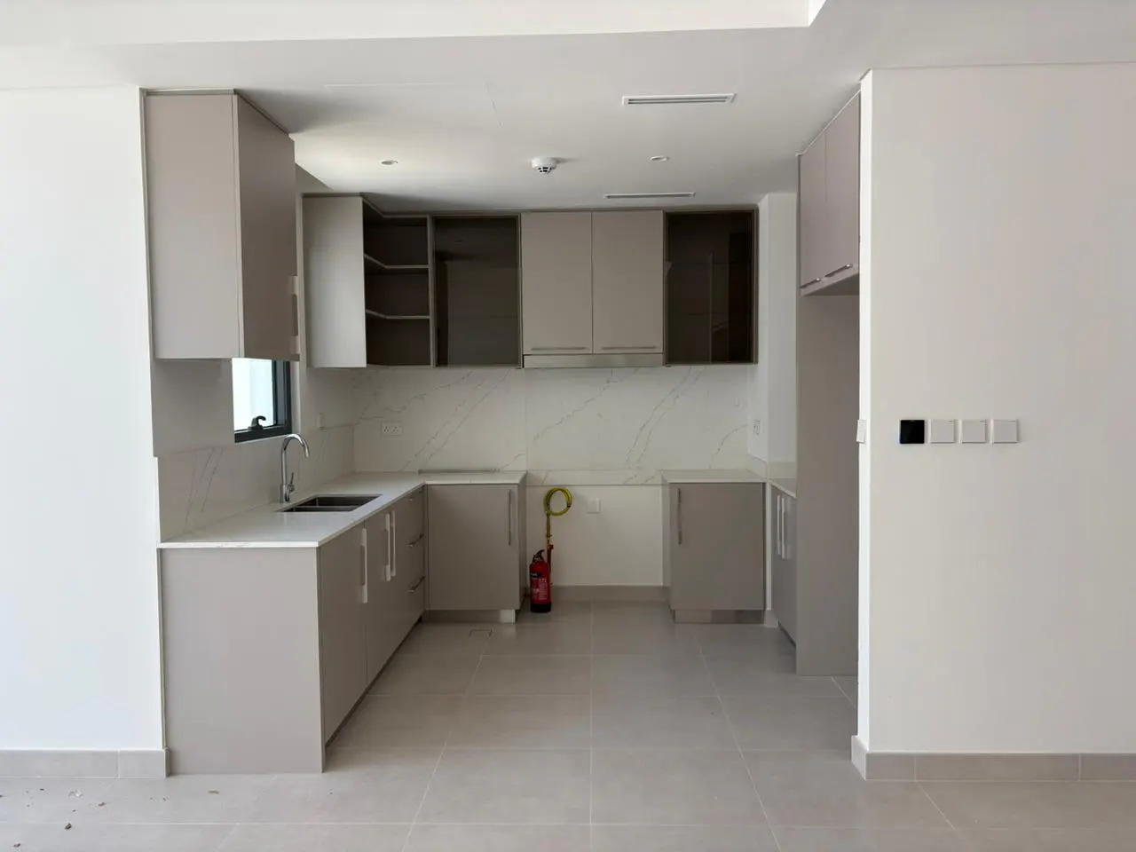 A modern kitchen with gray cabinets, white countertops, and tile flooring. A red fire extinguisher is visible near the cabinets.