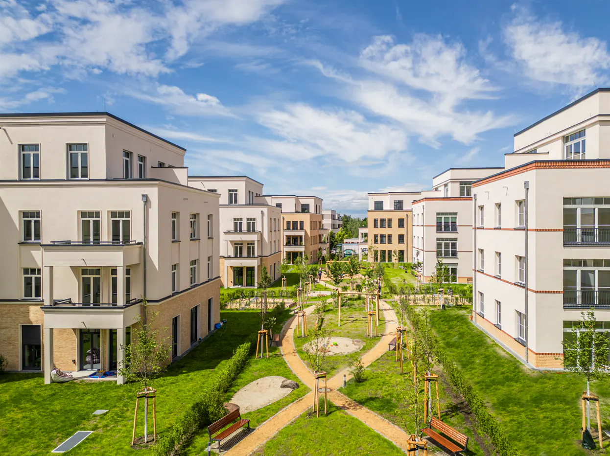 Apartment complex with a green courtyard. Beige buildings with dark windows surround a lawn with benches and winding paths. Blue sky above.