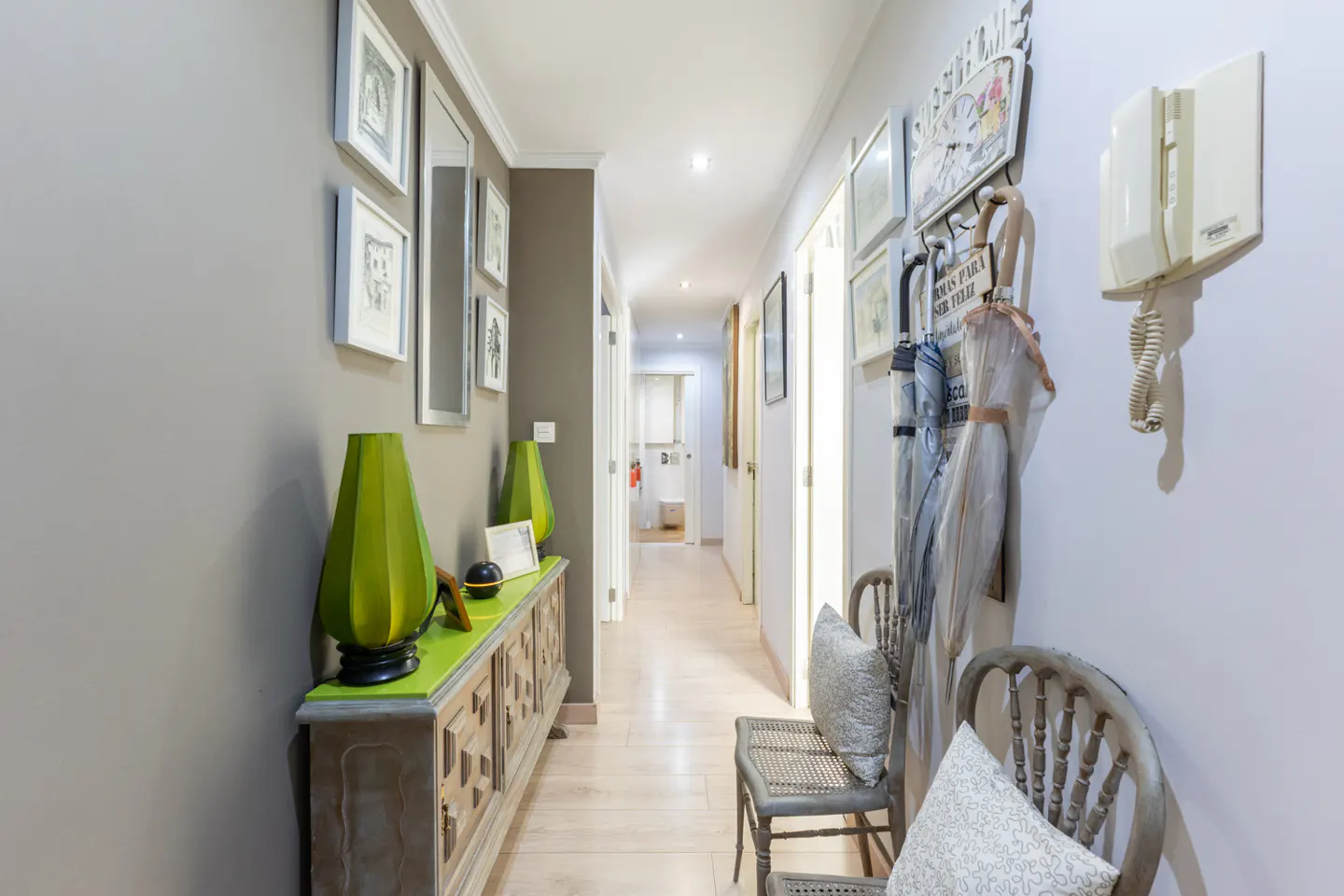 Hallway with gray walls, light wood floors, and white trim. A green-topped cabinet holds lamps. Chairs and umbrellas line the right wall.