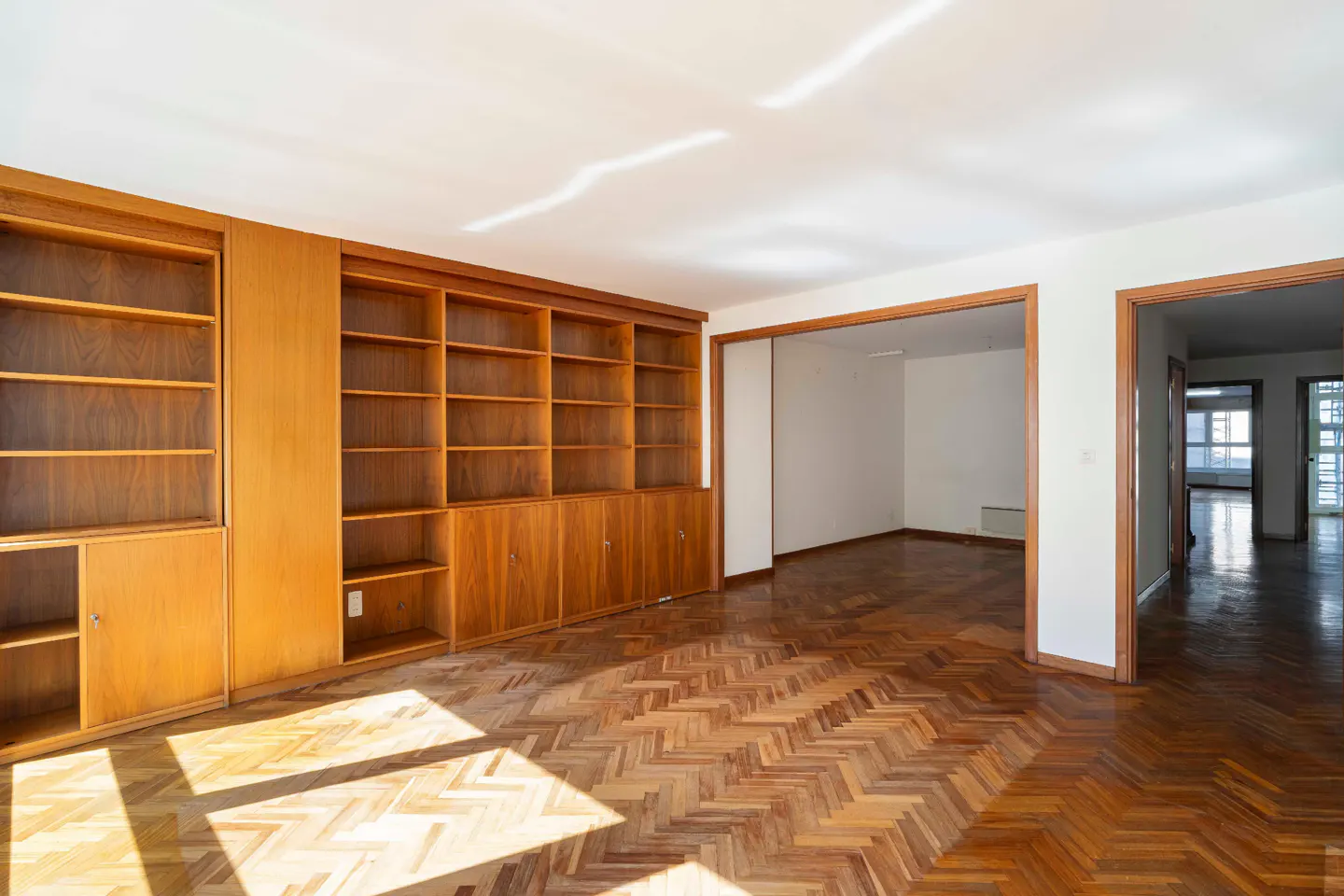 An empty living room with herringbone wood floors, built-in wood bookshelves, and doorways to other rooms.