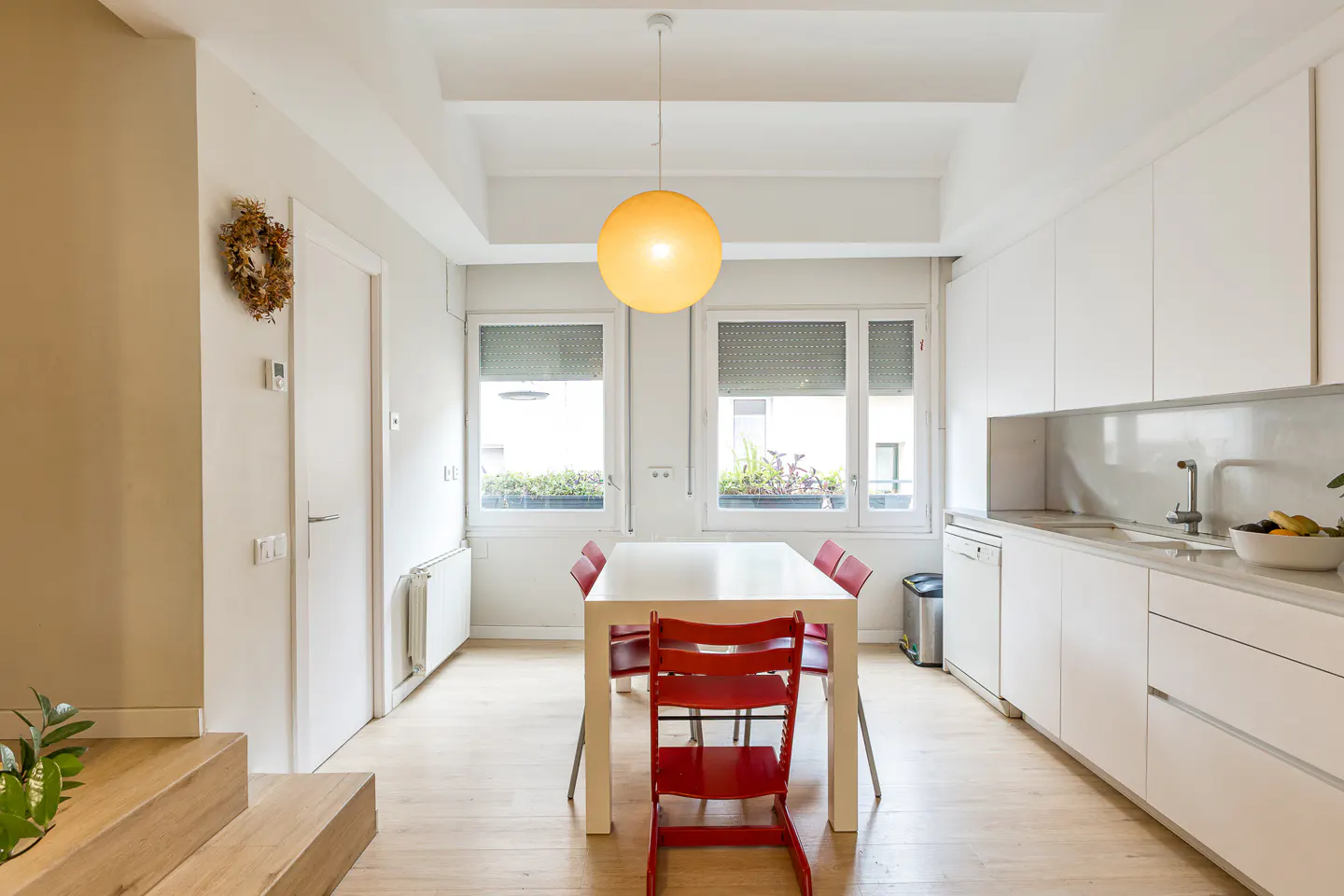 Bright, modern kitchen with white cabinets, a white table with red chairs, and a large, round, yellow pendant light. Windows overlook greenery.