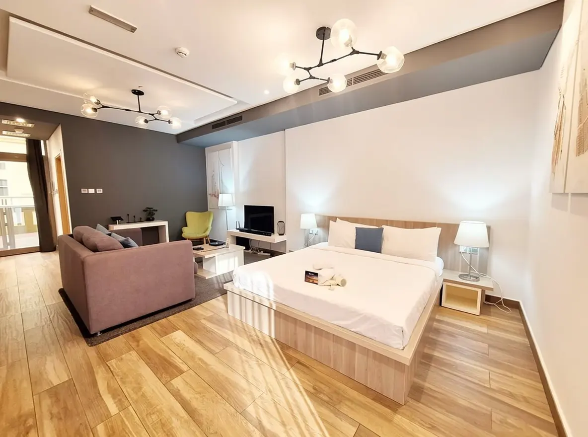 Bright studio apartment with a queen bed, mauve sofa, and a lime green chair. Modern light fixtures and wood floors.