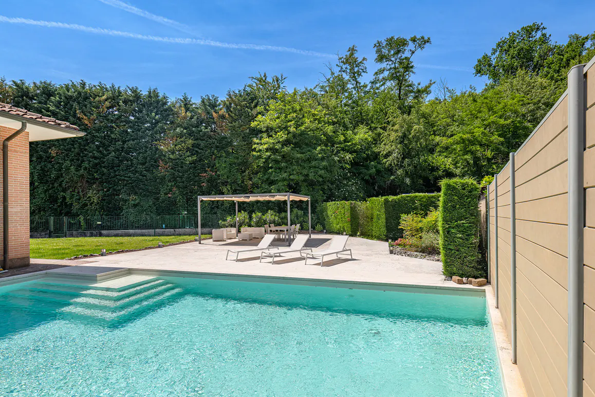 Backyard swimming pool with lounge chairs, a shaded patio area, and lush green trees under a clear blue sky.