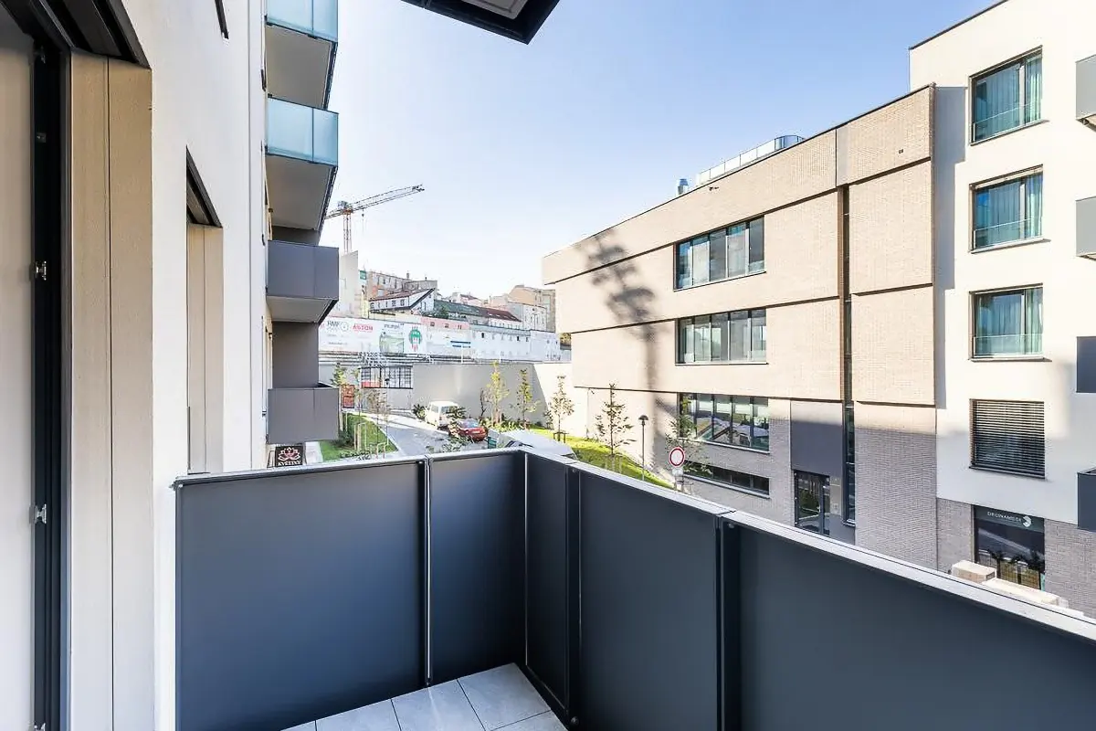 View from a gray balcony overlooking a modern building, street, and distant cityscape under a clear sky.