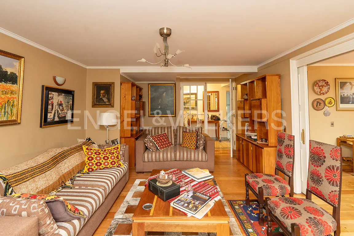 Living room with striped sofas, wooden bookshelves, and a coffee table with books. Paintings adorn the walls. Two chairs with floral patterns are visible.
