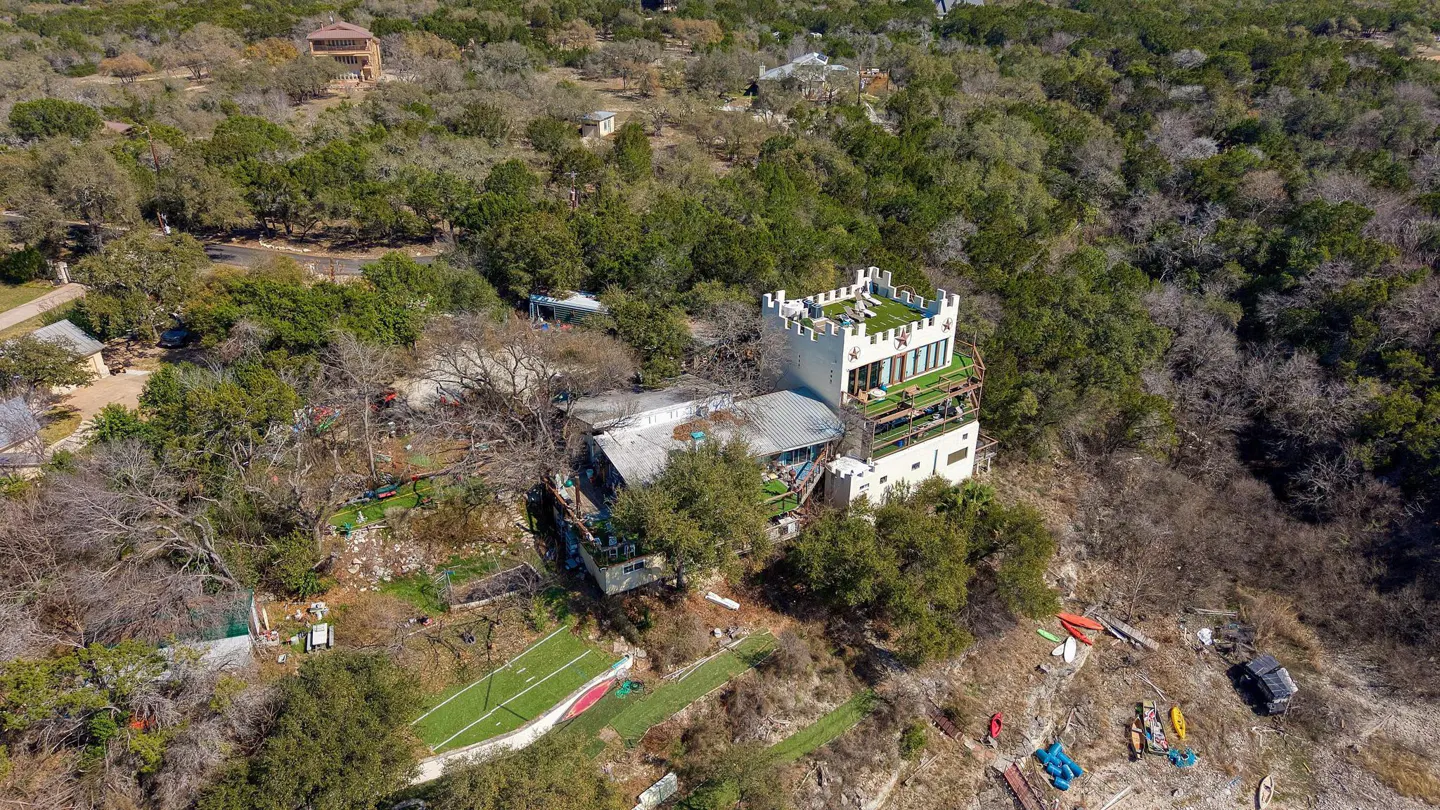 Aerial view of a white castle-like house surrounded by trees, with a green rooftop terrace and a sports field nearby.