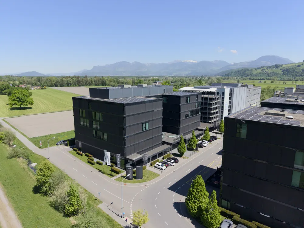 Aerial view of modern, dark-colored office buildings with cars parked outside. Green fields and mountains are visible in the background.