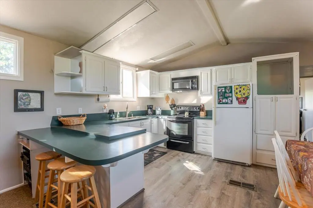 Bright kitchen with white cabinets, green countertops, and wood-look floors. Black appliances and a breakfast bar with stools are visible.