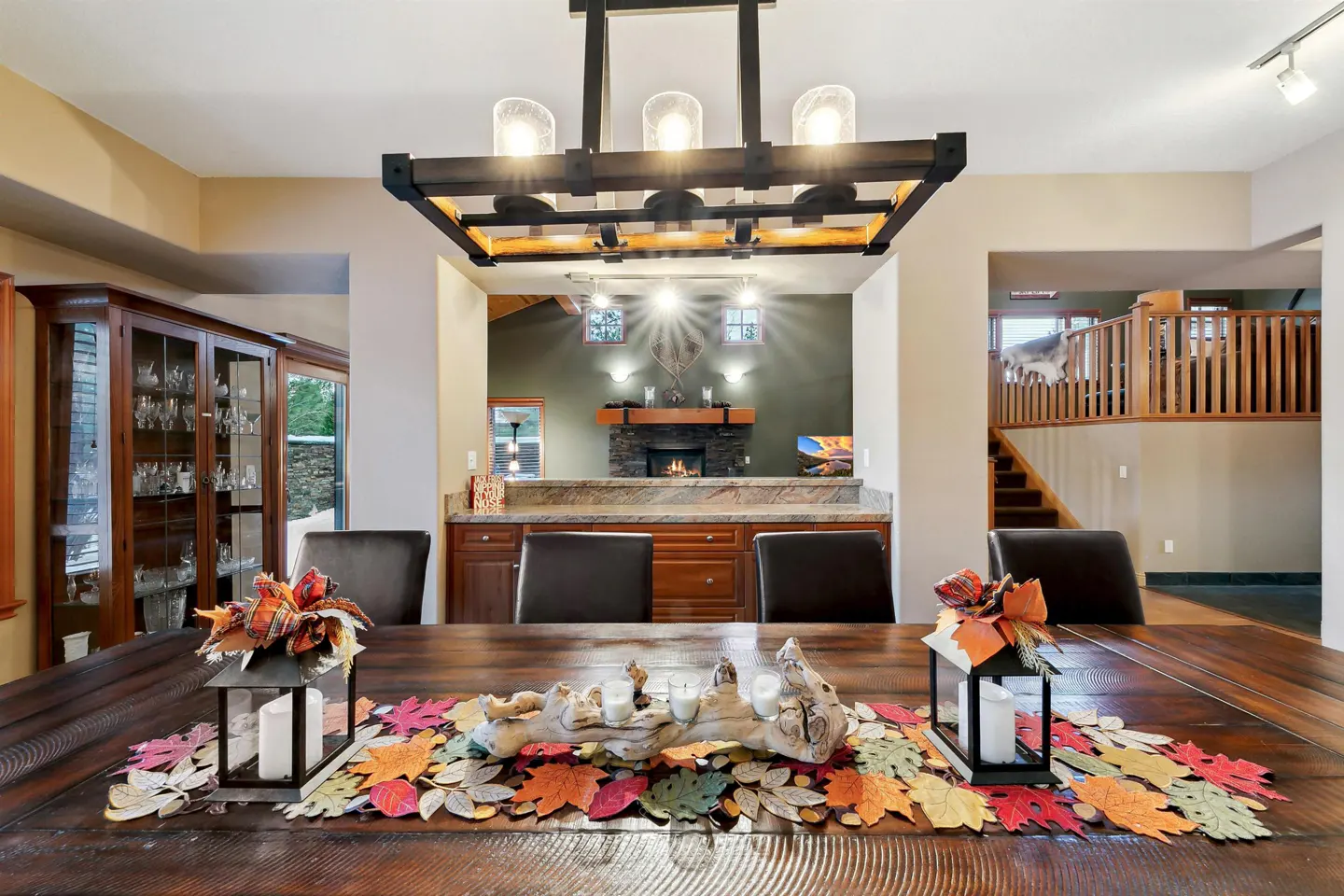 A dining room with a long wooden table decorated with fall leaves and lanterns. A modern chandelier hangs above, and a fireplace is visible in the background.