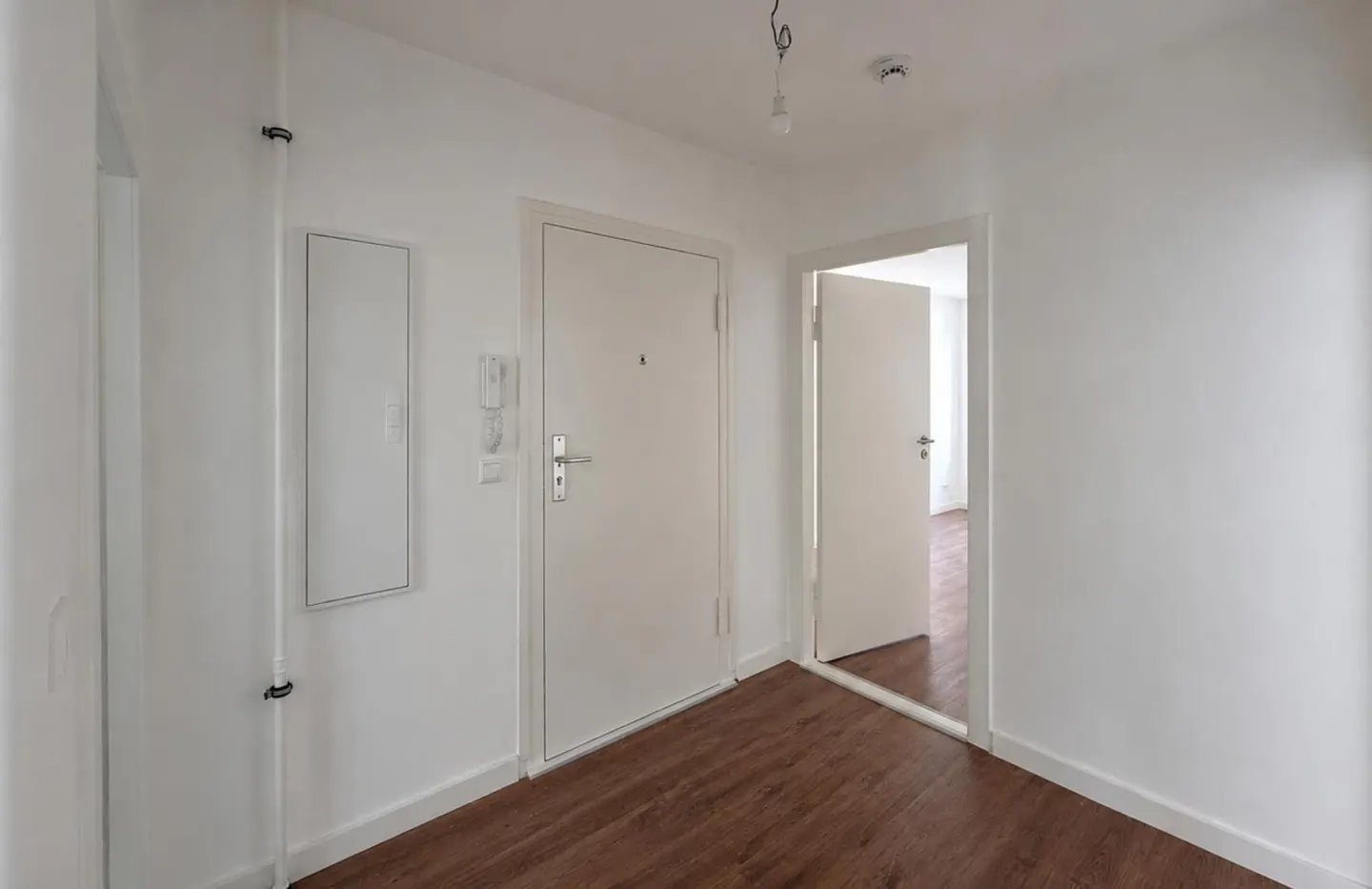 A bright hallway with white walls, wood-look flooring, a closed white door, and an open doorway leading to another room.