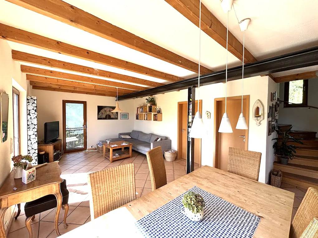 Open-plan living room with wooden beams, tiled floor, dining table with wicker chairs, and a gray sofa in the background.