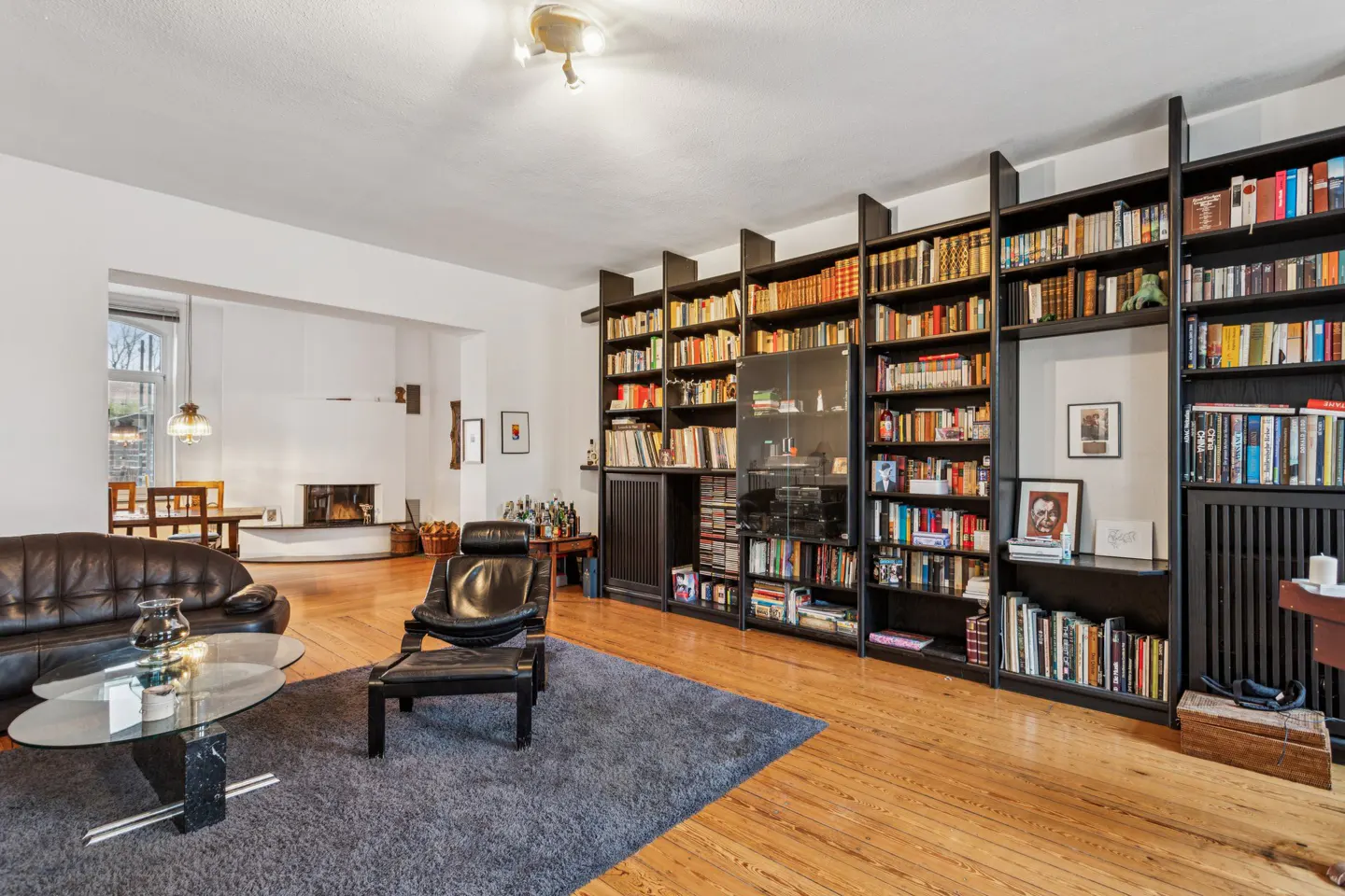 A living room with hardwood floors, a gray rug, a black leather chair, and a large black bookcase filled with books.