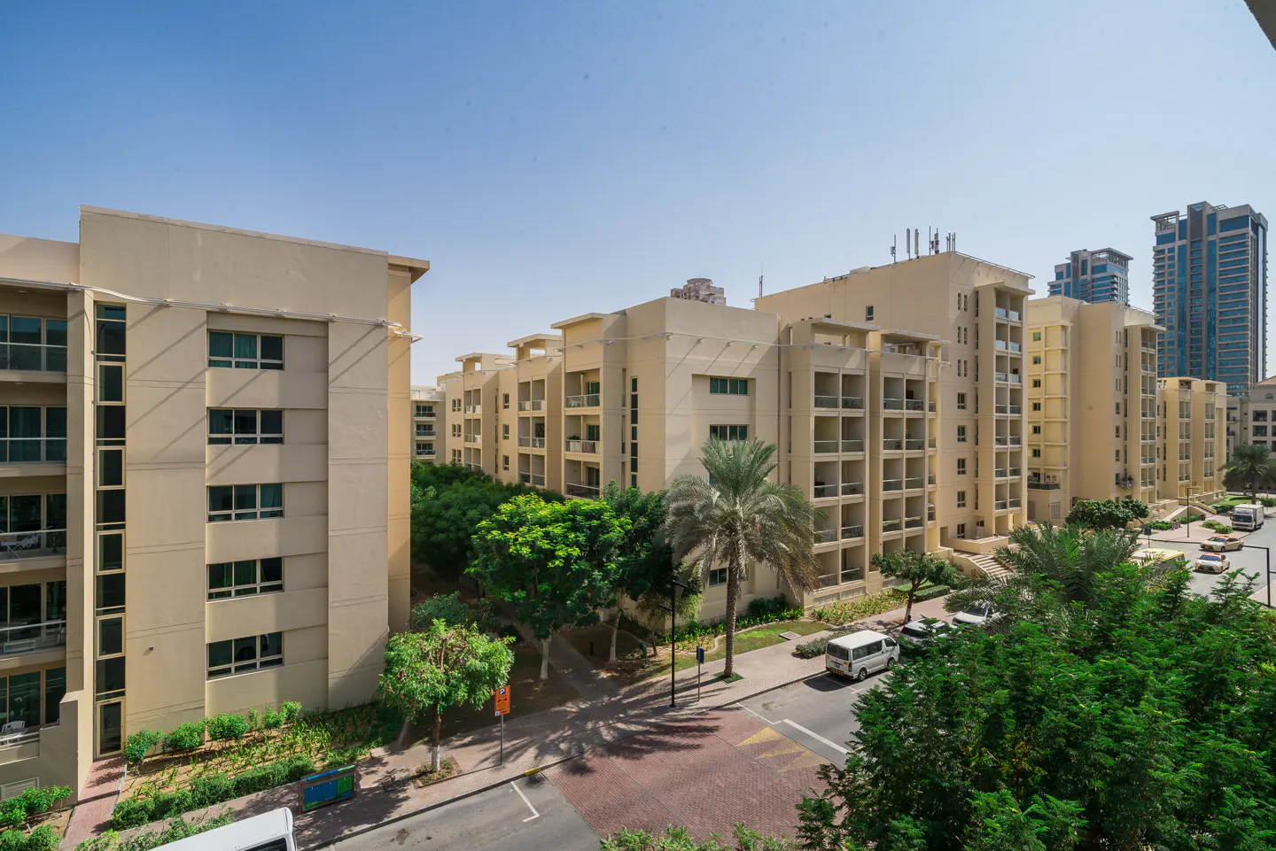 Exterior view of beige apartment buildings with balconies, green trees, and a red brick road under a clear blue sky.