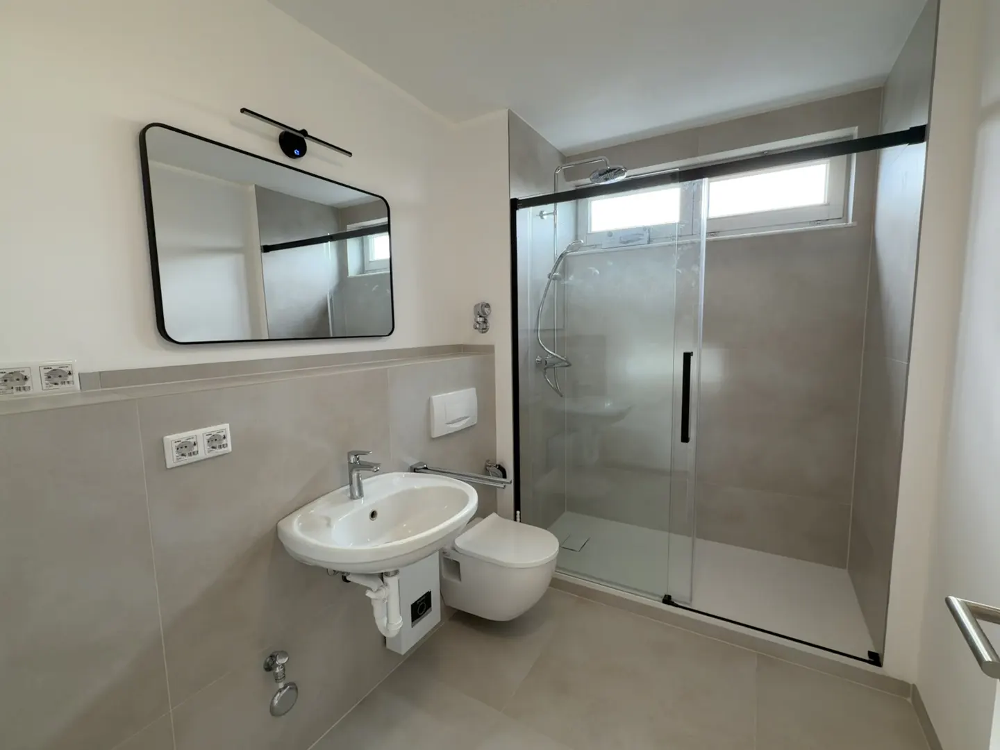 Modern bathroom with a white sink, toilet, and glass-enclosed shower. A black-framed mirror hangs above the sink. Gray tiles cover the walls and floor.