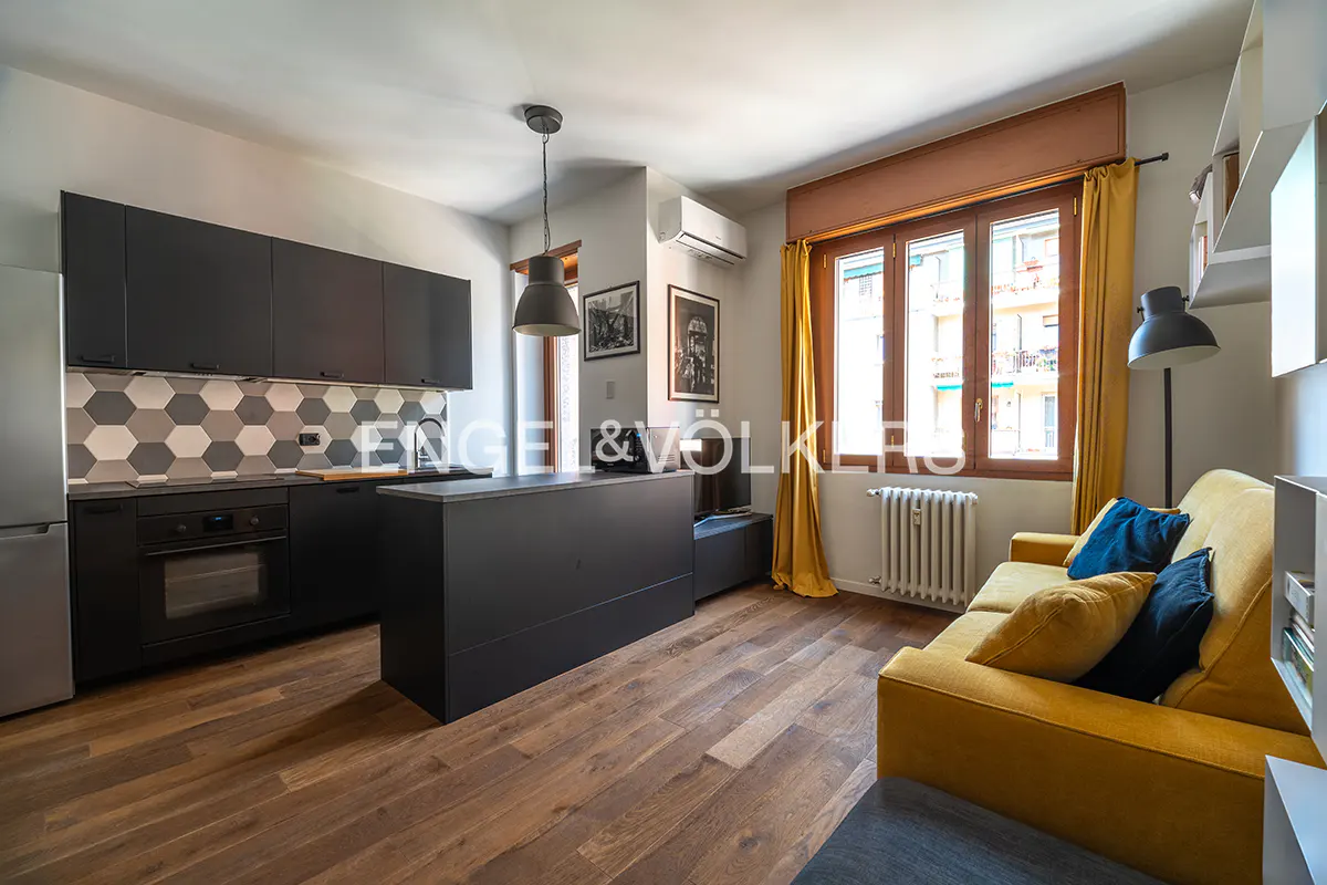 A modern apartment interior with a gray kitchen, island, and yellow sofa with blue pillows. A window with yellow curtains is in the background.