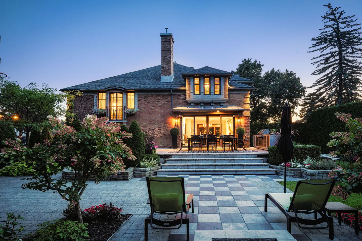 Brick house with a stone patio at dusk. Outdoor dining set and lounge chairs are visible.