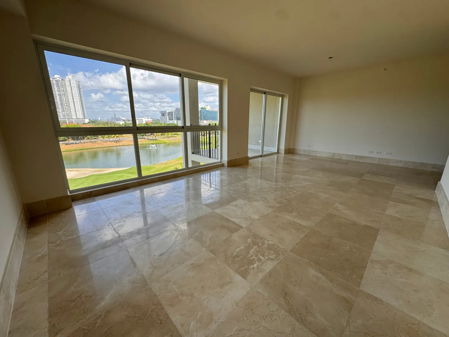 Empty room with beige marble floors and white walls. Large window overlooks a lake, golf course, and city skyline. Sliding glass doors to a balcony.