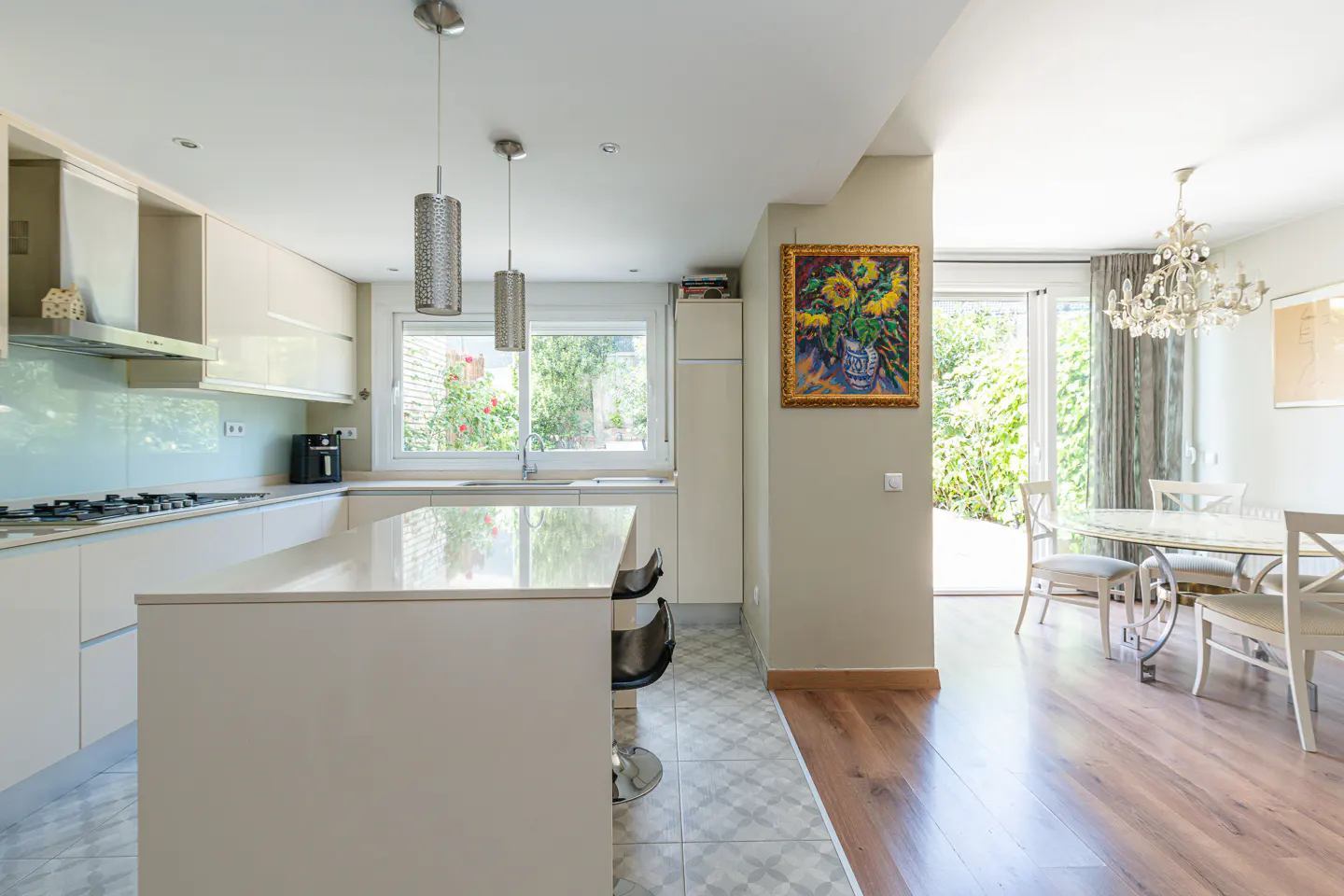Bright, modern kitchen with white cabinets, island with stools, and stainless steel pendant lights. Dining area with chandelier and garden view.