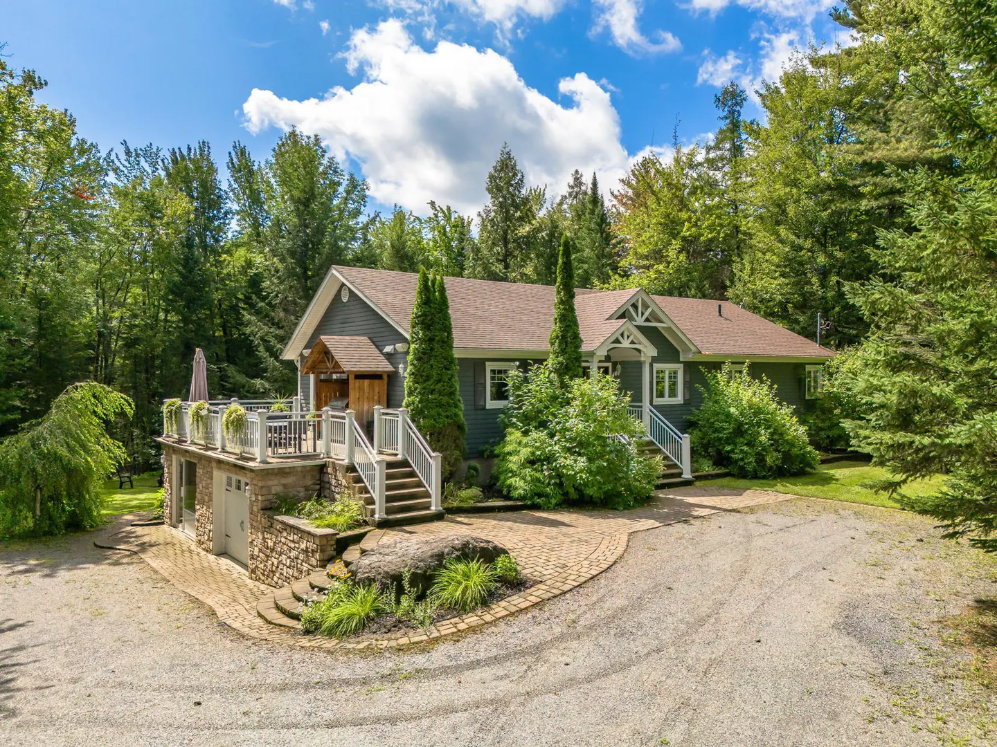 Exterior of a gray house with a brown roof, surrounded by trees and a gravel driveway. A stone patio is on the left.