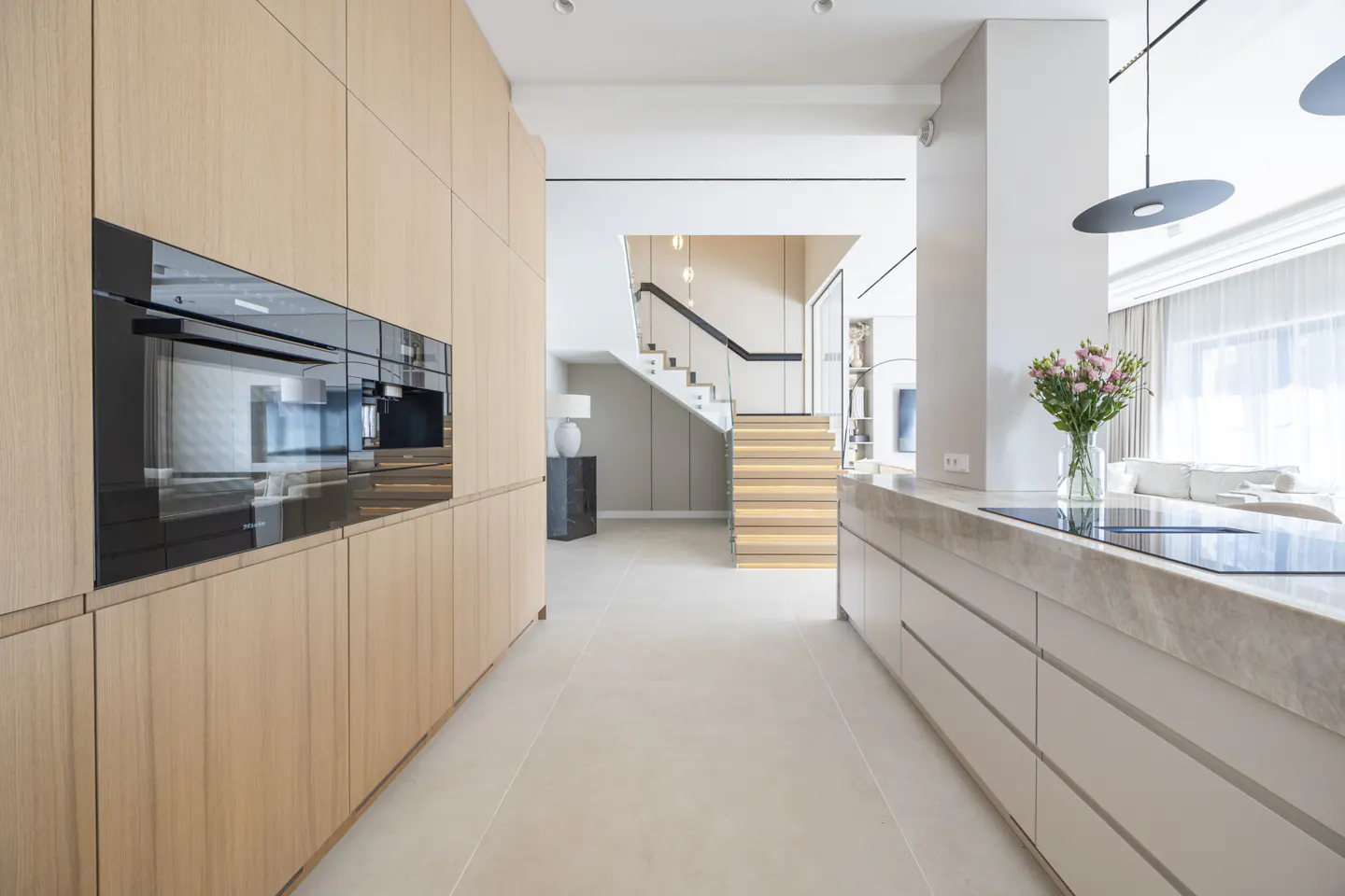 Modern kitchen with light wood cabinets, black appliances, and a marble countertop island. A staircase leads to the upper level.