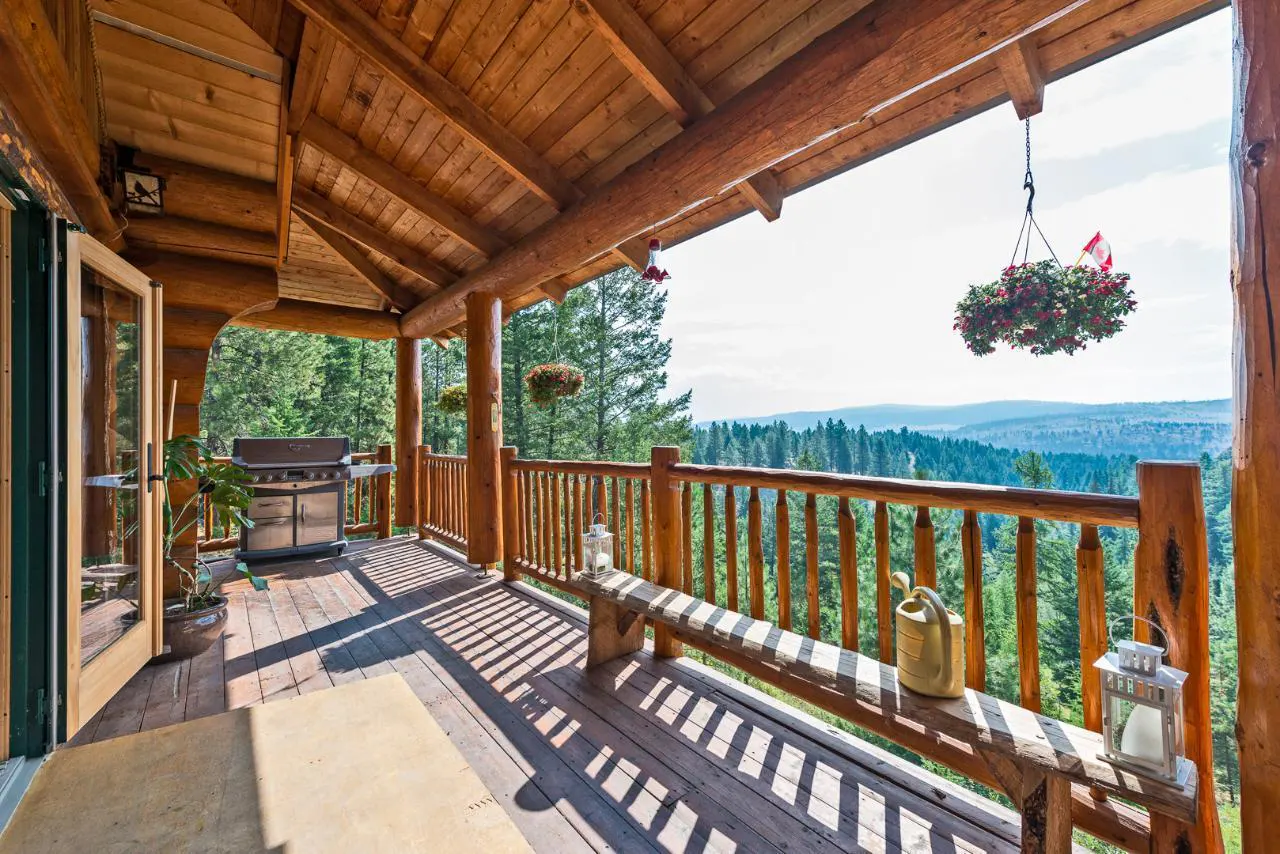 Log cabin porch with wood railing overlooking a forest. Bench, grill, and hanging flower baskets decorate the space.
