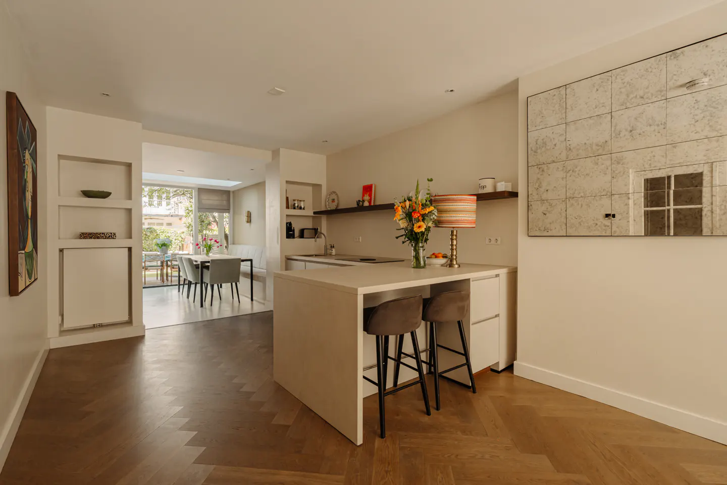 A bright, modern kitchen with a white island, two stools, and a vase of flowers. A dining area is visible in the background.